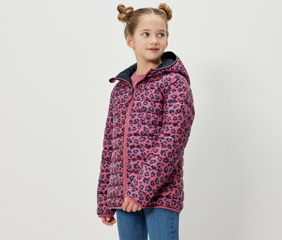 Une fille pose en veste softshell et matelassée 2 en 1 pour enfants. La veste a un fond rose avec un motif animal bleu marine.