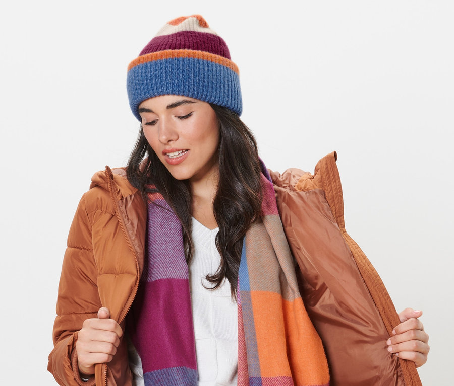 Une femme en veste marron ajuste son bonnet en maille et porte une écharpe.