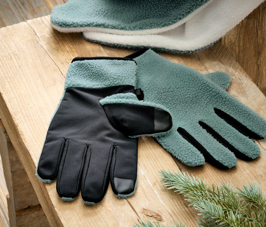 Une paire de gants turquoise et noirs en laine polaire reposent sur une table en bois à côté d'une branche de sapin.