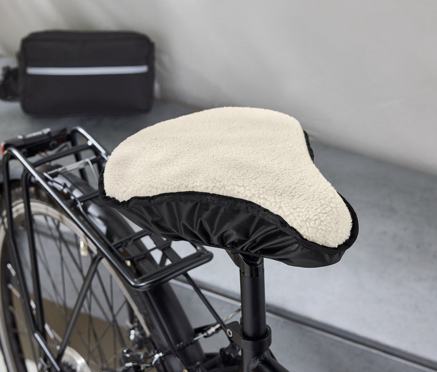 Gros plan d'un vélo avec un cadre noir, dont la selle est recouverte d'une housse beige et duveteuse. Un sac noir avec une bande blanche est visible à l'arrière-plan.