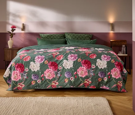 Un lit avec un couvre-lit matelassé vert foncé orné de grandes fleurs roses, rouges et blanches. Quatre oreillers verts sont à la tête du lit, flanqués de tables de chevet en bois et d'un mur bicolore.