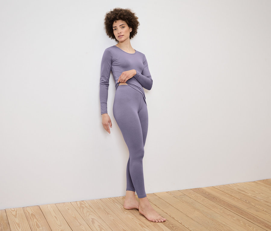 Une femme pose contre un mur dans un ensemble lilas composé d'un haut à manches longues et d'un legging.