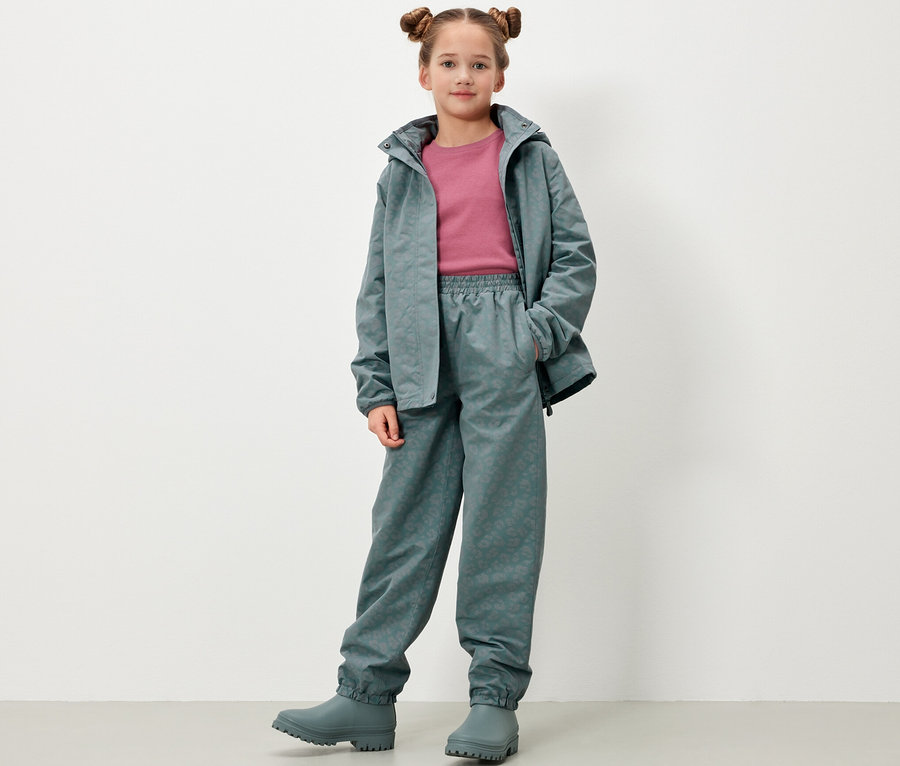 Une fille pose, portant un pantalon de pluie pour enfants avec des éléments réfléchissants, une veste et des bottes en caoutchouc gris-vert.