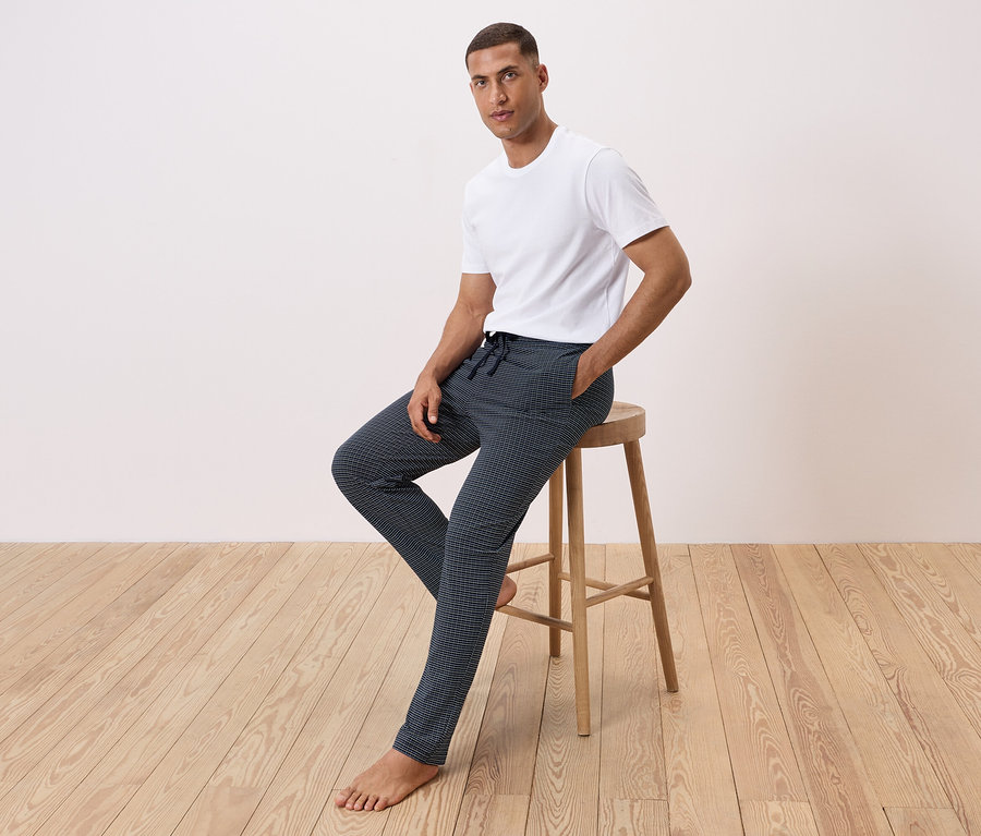 Un homme est assis sur un tabouret, portant un t-shirt blanc et un pantalon à motifs.