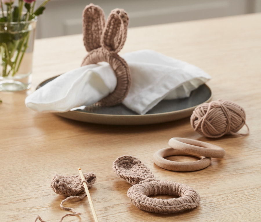 Sur une table se trouve un kit crochet DIY « ronds de serviette ». Un rond fini avec des oreilles de lapin est posé sur une serviette dans une assiette. À côté se trouvent un ouvrage au crochet en cours, une pelote de laine et des anneaux en bois.