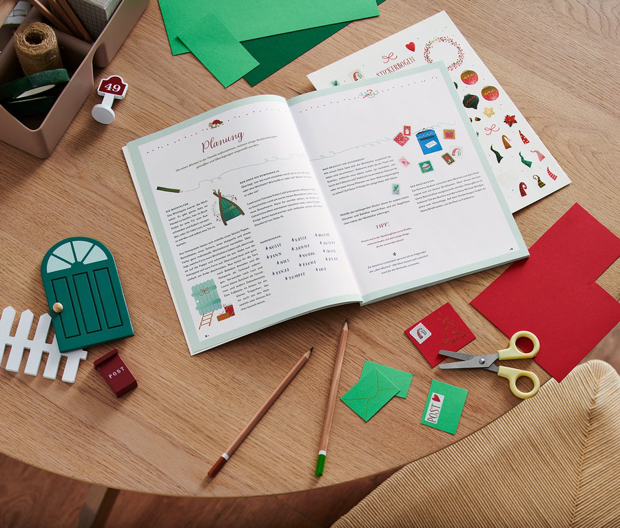 Sur une table en bois sont posés un livre ouvert, des ciseaux, des crayons de couleur, des papiers colorés et des décorations, dont le livre « Hurra, ein Wichtel zieht ein! » (en allemand).