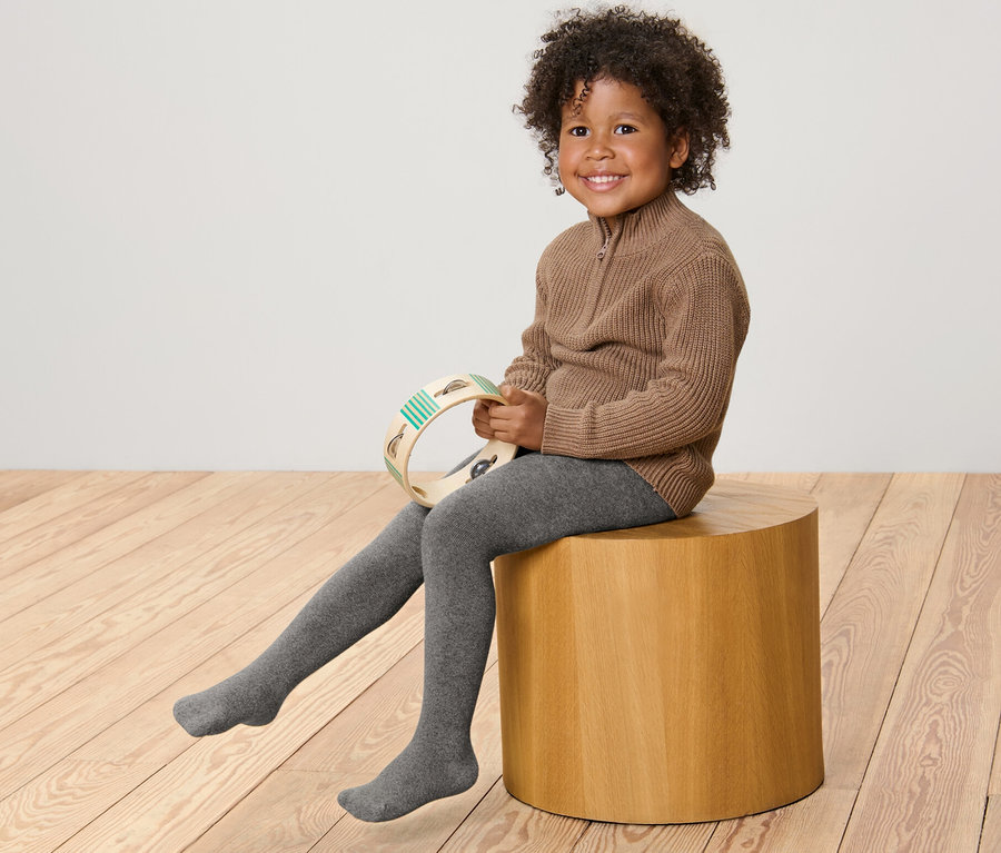 Un enfant souriant est assis sur un tabouret en bois et tient un tambourin. Il porte un pull marron et 2 collants thermiques pour enfant chinés gris.