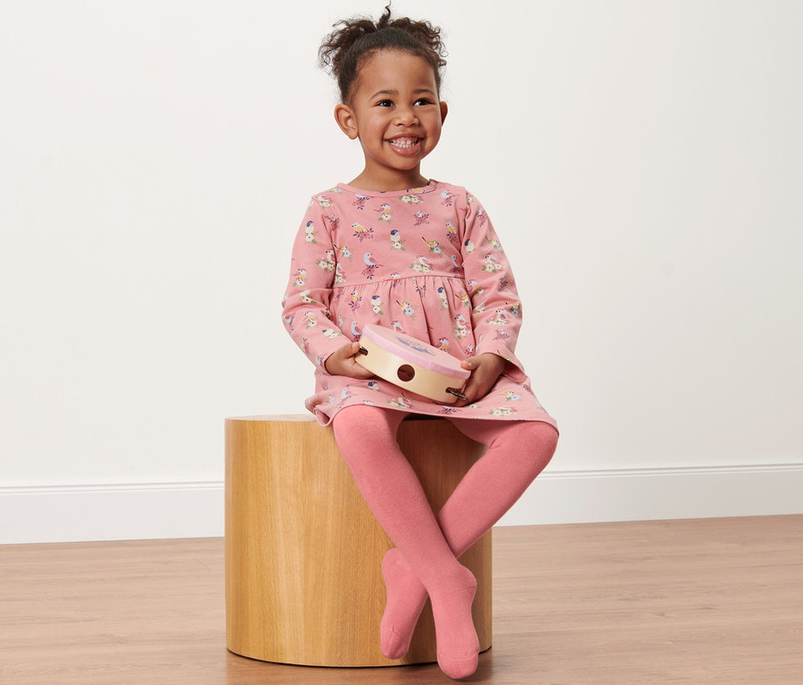 Petite fille souriante assise sur un tabouret en bois tenant un tambourin. Elle porte une robe rose imprimée et 2 collants thermiques pour tout-petit, oiseau.