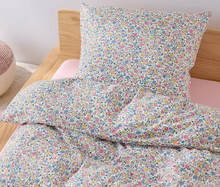 Sur un lit se trouve une parure de lit pour enfant en renforcé de taille normale avec un motif floral, à côté se trouve un pouf rose.