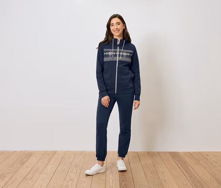 Une femme se tient sur un plancher en bois et porte un sweat à capuche zippé bleu marine, un pantalon de survêtement et des baskets blanches.