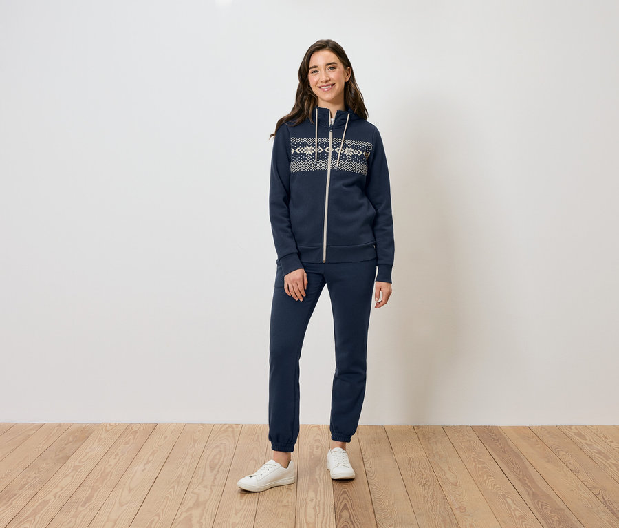 Une femme se tient sur un plancher en bois et porte un sweat à capuche zippé bleu marine, un pantalon de survêtement et des baskets blanches.