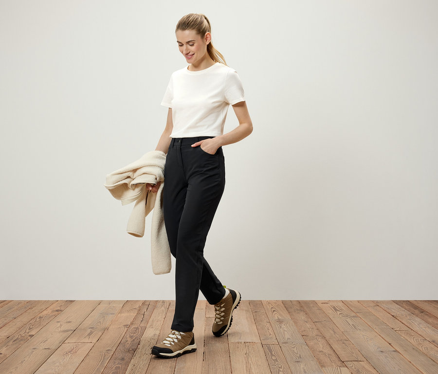 Une femme marche sur un plancher en bois, portant un pantalon noir, un t-shirt blanc et des chaussures marron. Elle tient un pull crème à la main.