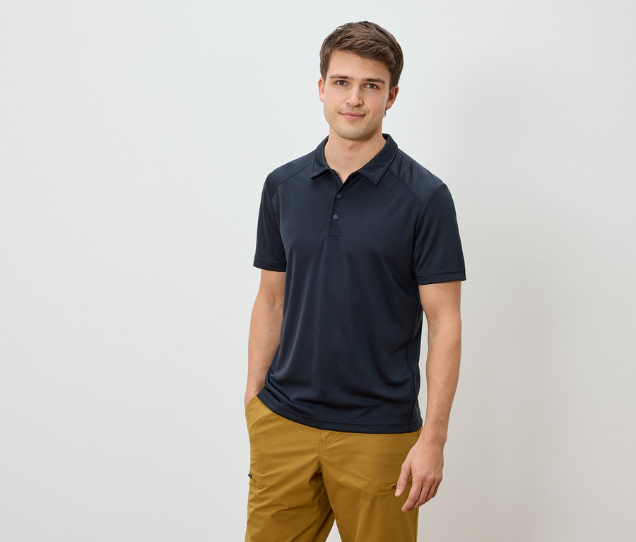 Homme portant un polo bleu marine et un pantalon marron doré, une main dans la poche.