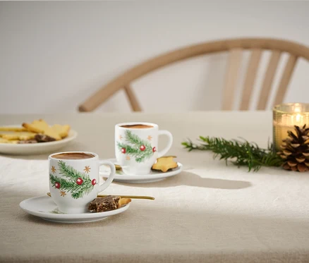 Deux tasses à expresso avec un motif de Noël sur une table avec des biscuits et des bougies.
