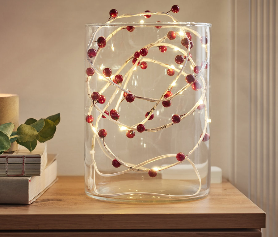 Une branche décorative à LED avec des baies rouges est placée dans un vase en verre sur une commode en bois.