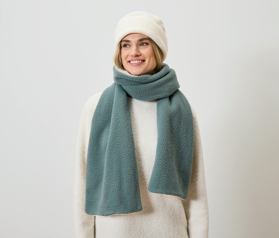 Une femme blonde souriante porte un pull crème, une écharpe bleu-vert et un bonnet crème.