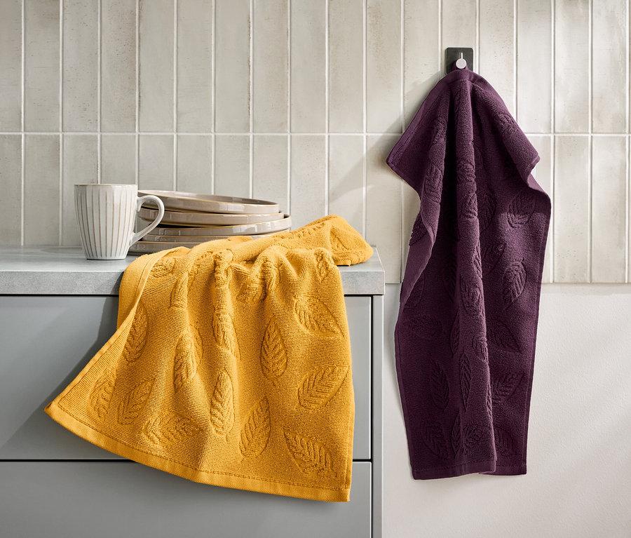 Une serviette jaune avec un motif de feuilles repose sur une armoire grise à côté d'une tasse et d'assiettes. Une serviette violette est suspendue à un crochet au mur.