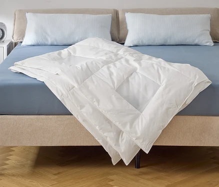 Une couette blanche est posée sur un lit avec un drap-housse bleu et deux oreillers. Un ventilateur est posé sur la table de chevet.