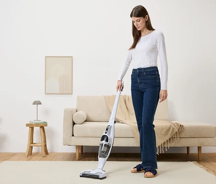 Une femme passe l'aspirateur sur un tapis beige dans un salon avec un aspirateur sans fil blanc.