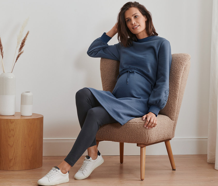 Une femme enceinte est assise dans un fauteuil et porte une robe molletonnée de grossesse et d'allaitement, des leggings et des baskets.