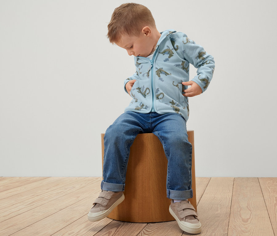 Un garçon est assis sur un tabouret et porte une veste polaire pour enfant avec un motif de dragon.