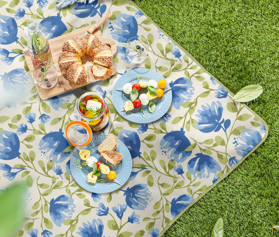 Sur l'herbe, une petite nappe de pique-nique à motif floral. Des assiettes de nourriture, des verres de boissons et une planche à découper en bois avec des pâtisseries sont disposés dessus.