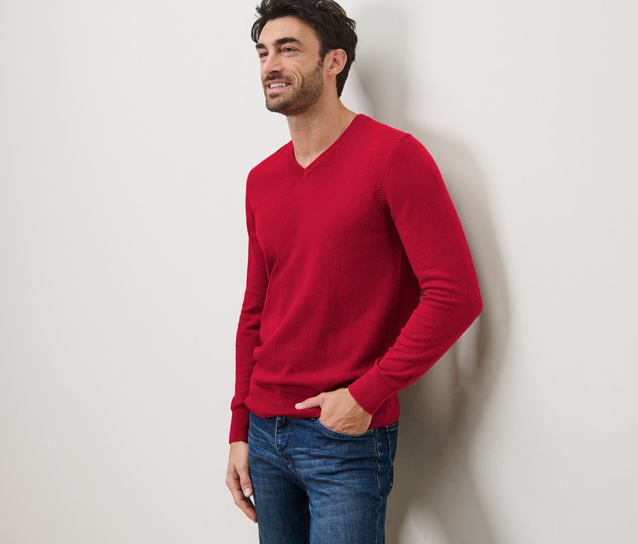 Un homme souriant est appuyé contre un mur et porte un pull rouge à col en V et un jean.