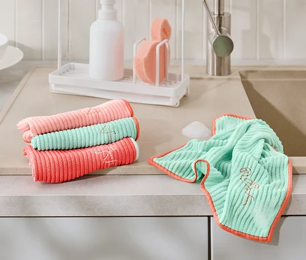 Sur un comptoir de cuisine reposent, pliés et dépliés, 4 chiffons de nettoyage universels corail, rose et turquoise avec broderie flamant rose.