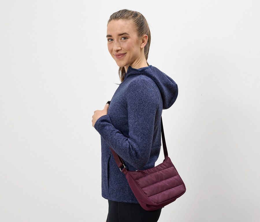 Une femme aux cheveux bruns et au sweat à capuche bleu sourit par-dessus son épaule. Elle porte un sac à main violet.