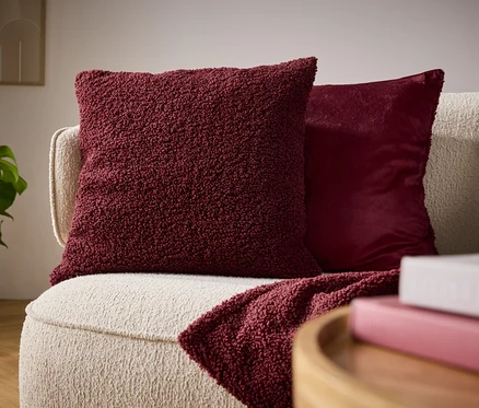Gros plan d'un canapé beige avec un coussin en peluche bordeaux, un coussin brillant et un plaid drapé.