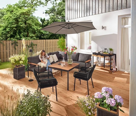 Sur une terrasse, une femme est assise avec un chien sur un coin salon-salle à manger »Elin«. On peut également voir un chariot de grill avec un plan de travail en polyciment, un pot de fleurs avec un tressage textile, un lampadaire d'extérieur LED sur batterie, une lampe de table d'extérieur LED sur batterie, deux coussins d'extérieur à toucher lin et une chaise de jardin avec un tressage croisé textile.