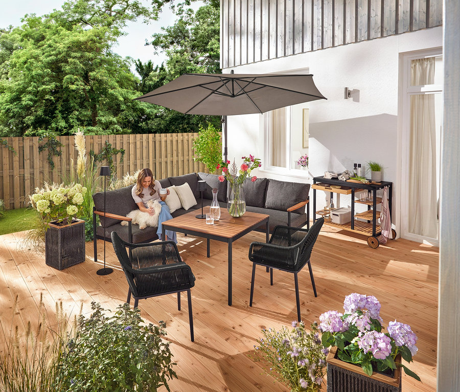 Sur une terrasse, une femme est assise avec un chien sur un coin salon-salle à manger »Elin«. On peut également voir un chariot de grill avec un plan de travail en polyciment, un pot de fleurs avec un tressage textile, un lampadaire d'extérieur LED sur batterie, une lampe de table d'extérieur LED sur batterie, deux coussins d'extérieur à toucher lin et une chaise de jardin avec un tressage croisé textile.