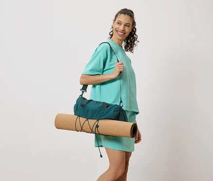Une femme souriante portant une tenue turquoise avec un sac de sport vert foncé et un tapis de yoga roulé sur l'épaule.
