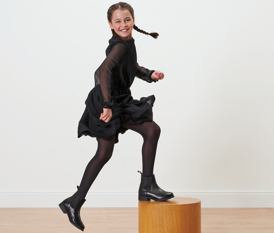 Fillette souriante avec des tresses en robe noire et 2 collants en nylon pour enfant noirs sautant d'un socle en bois.