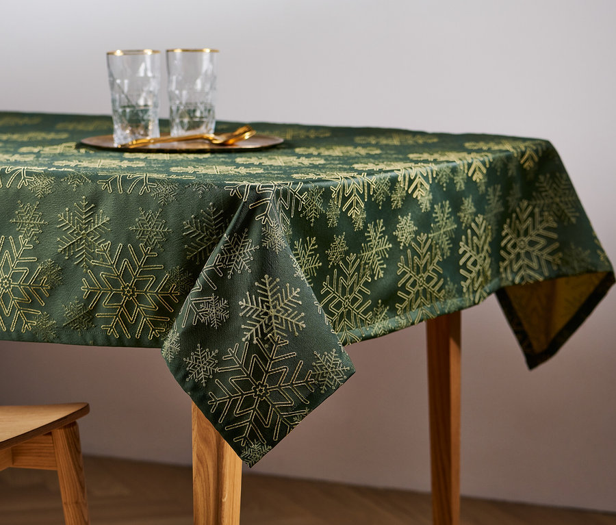 Sur une table en bois, une nappe jacquard verte avec un motif de flocons de neige. Sur la table, des verres et une chaise en bois.