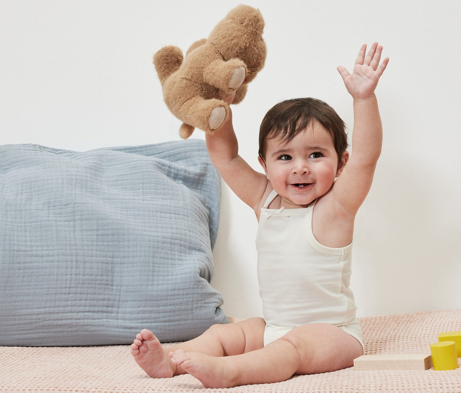 Un bébé souriant est assis et tient un ours en peluche. Il porte 5 bodys pour bébé.