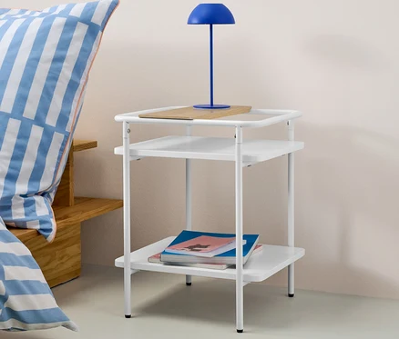 Table d’appoint blanche avec deux étagères, une planche en bois sur l’étagère supérieure et une lampe bleue dessus. Des livres sont posés sur l’étagère inférieure.