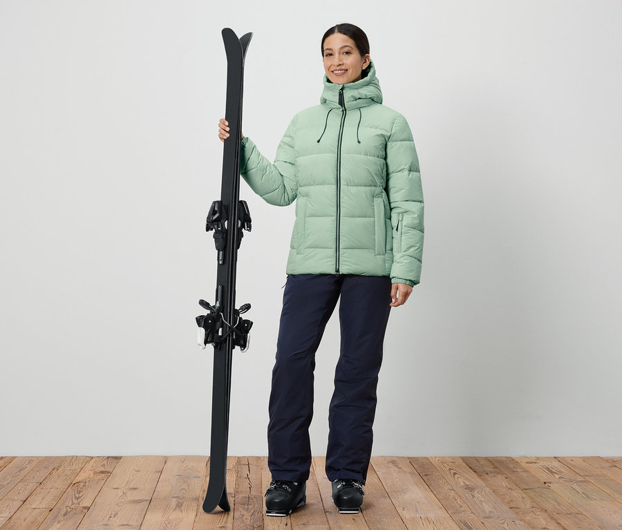 Femme tenant des skis, portant une veste verte et un pantalon bleu marine.