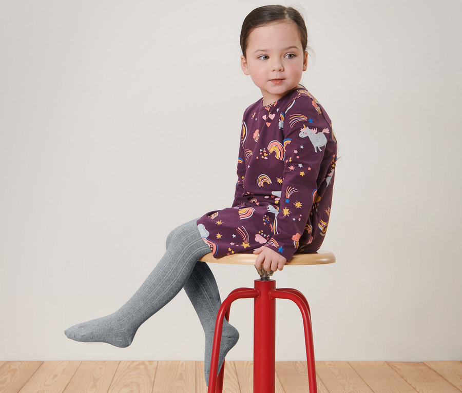 Une fille est assise sur un tabouret rouge et porte une robe imprimée et 2 paires de collants pour enfant gris.