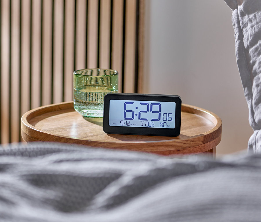 Réveil radio numérique à écran LCD sur une table de chevet à côté d'un lit.