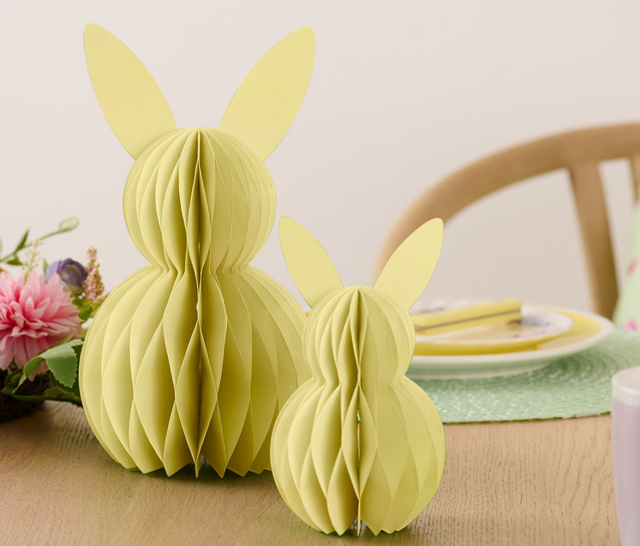 Deux lapins de Pâques en papier sont posés sur une table à côté de fleurs et d'assiettes.