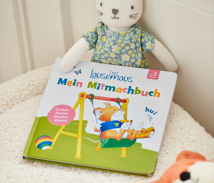 Livre « Leo Lausemaus – mein Mitmachbuch » avec une peluche de chat et d'autres jouets sur une surface duveteuse.