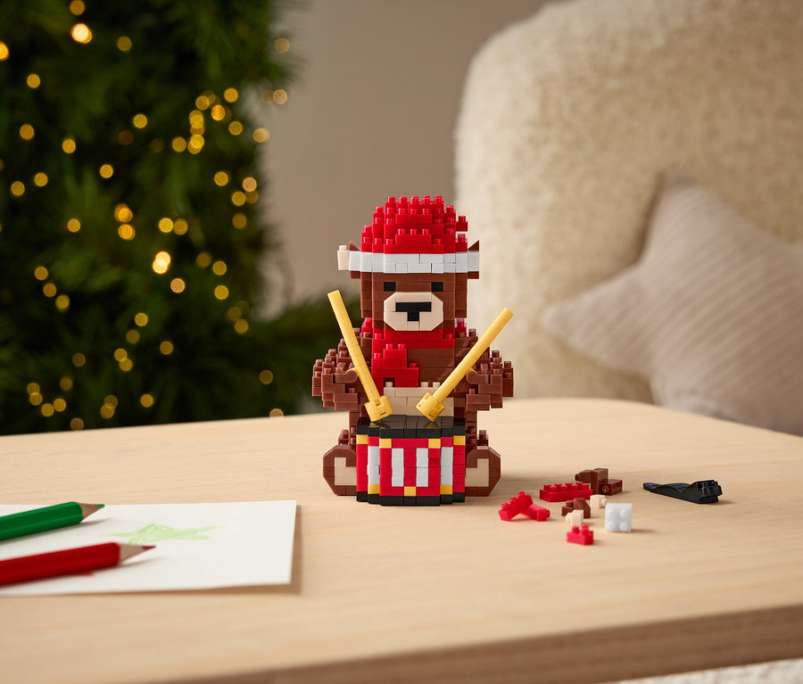 Un ours en forme de blocs avec un chapeau et une écharpe rouges joue du tambour avec des baguettes sur une table.