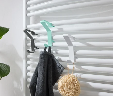 Des crochets noirs, verts et blancs sont accrochés à un radiateur blanc. Une serviette grise est accrochée au crochet vert et un luffa au crochet blanc.