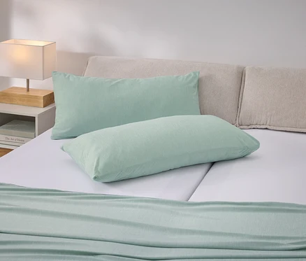 Lit avec coussins vert clair et couvre-lit sur des draps blancs. Table de chevet avec lampe à gauche.