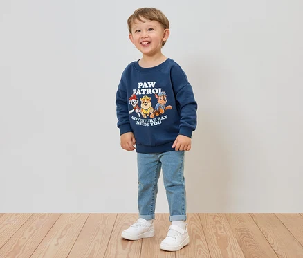 Un garçon souriant porte un sweat-shirt bleu Pat' Patrouille, un jean et des baskets blanches.