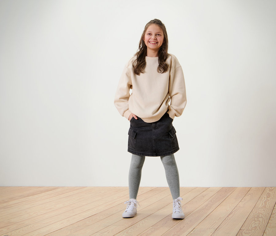 Une fille souriante pose en sweat-shirt beige, jupe noire et 5 paires de collants pour enfant gris.