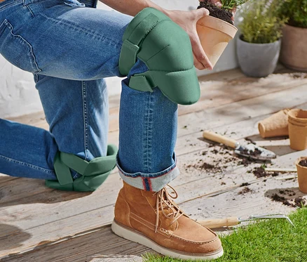 Une personne agenouillée portant des genouillères de jardinage tient un pot de fleurs contenant une plante.
