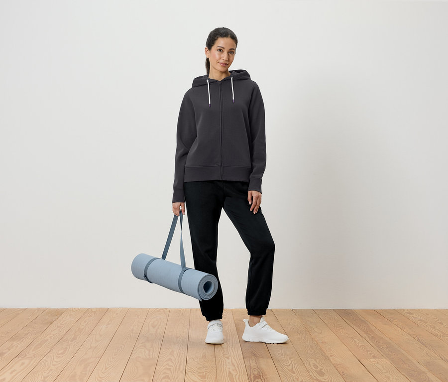 Femme tenant un tapis de yoga dans un sweat à capuche gris et un pantalon noir.