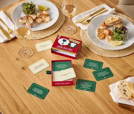 Sur une table en bois, deux assiettes contenant des crevettes, de la salade et du pain, deux verres de vin et un jeu Et toi donc ?